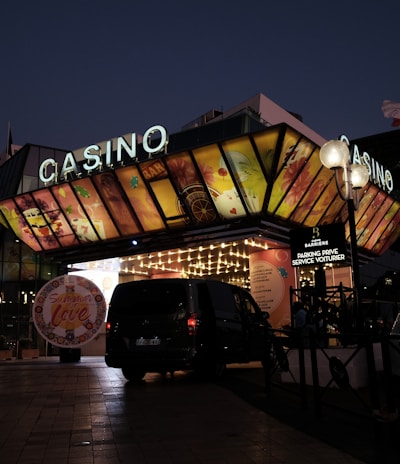 A building with a brightly illuminated entrance is seen at night. The sign above reads 'Casino' in bright letters. The facade features colorful graphics depicting various indulgent themes. Several vehicles are parked near the entrance, and people are visible in the vicinity. Tall lamp posts with spherical lights are present, casting a warm glow.